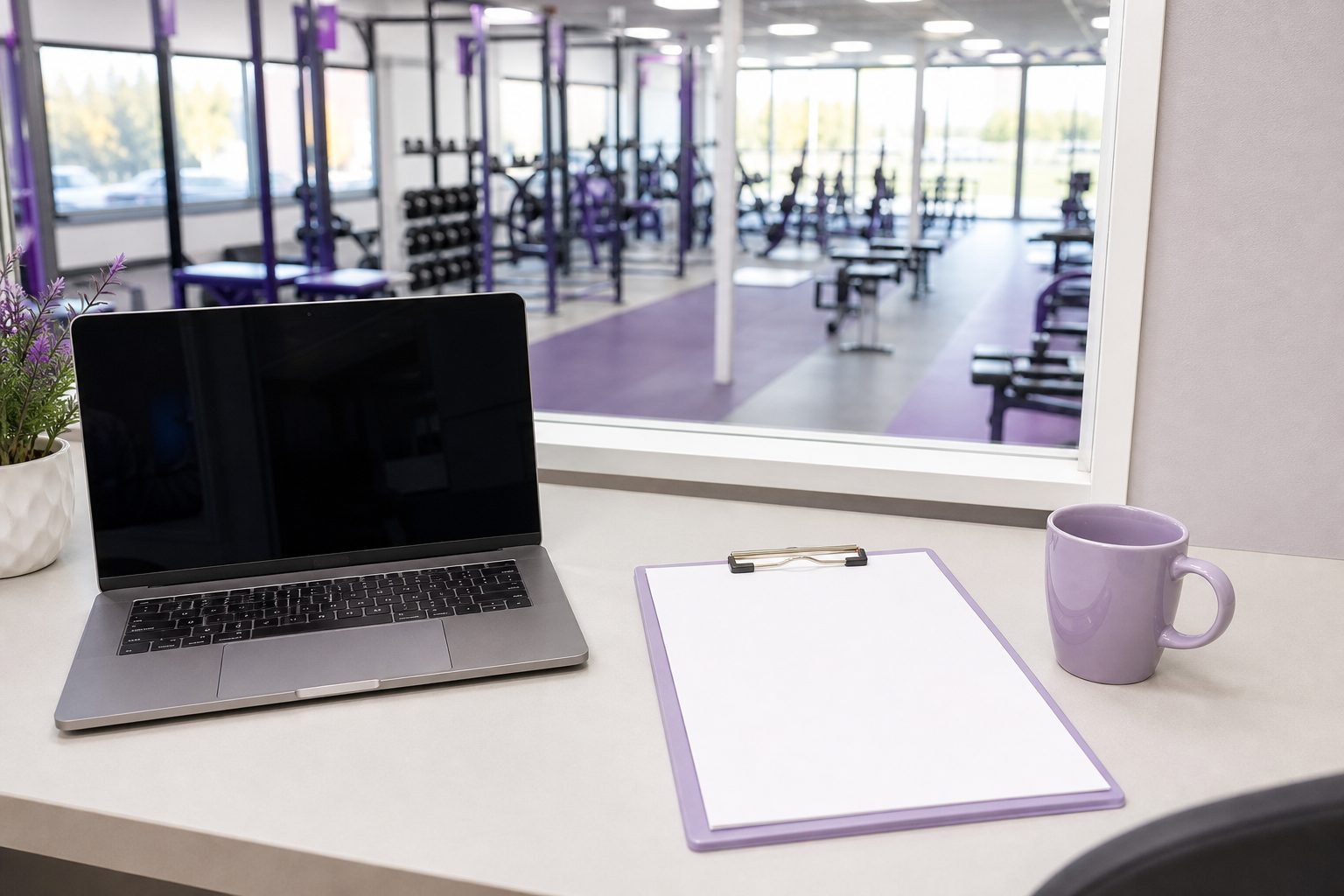 Gym office desk with laptop and clipboard beside a window over a bright training floor
