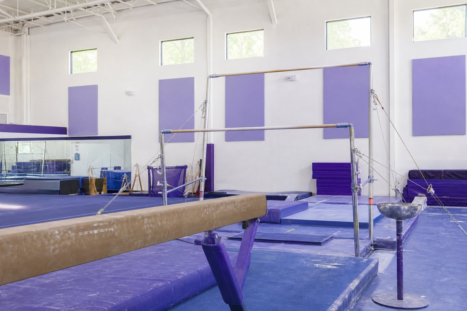 A bright gymnastics gym interior with a balance beam, uneven bars, blue landing mats and a chalk bowl